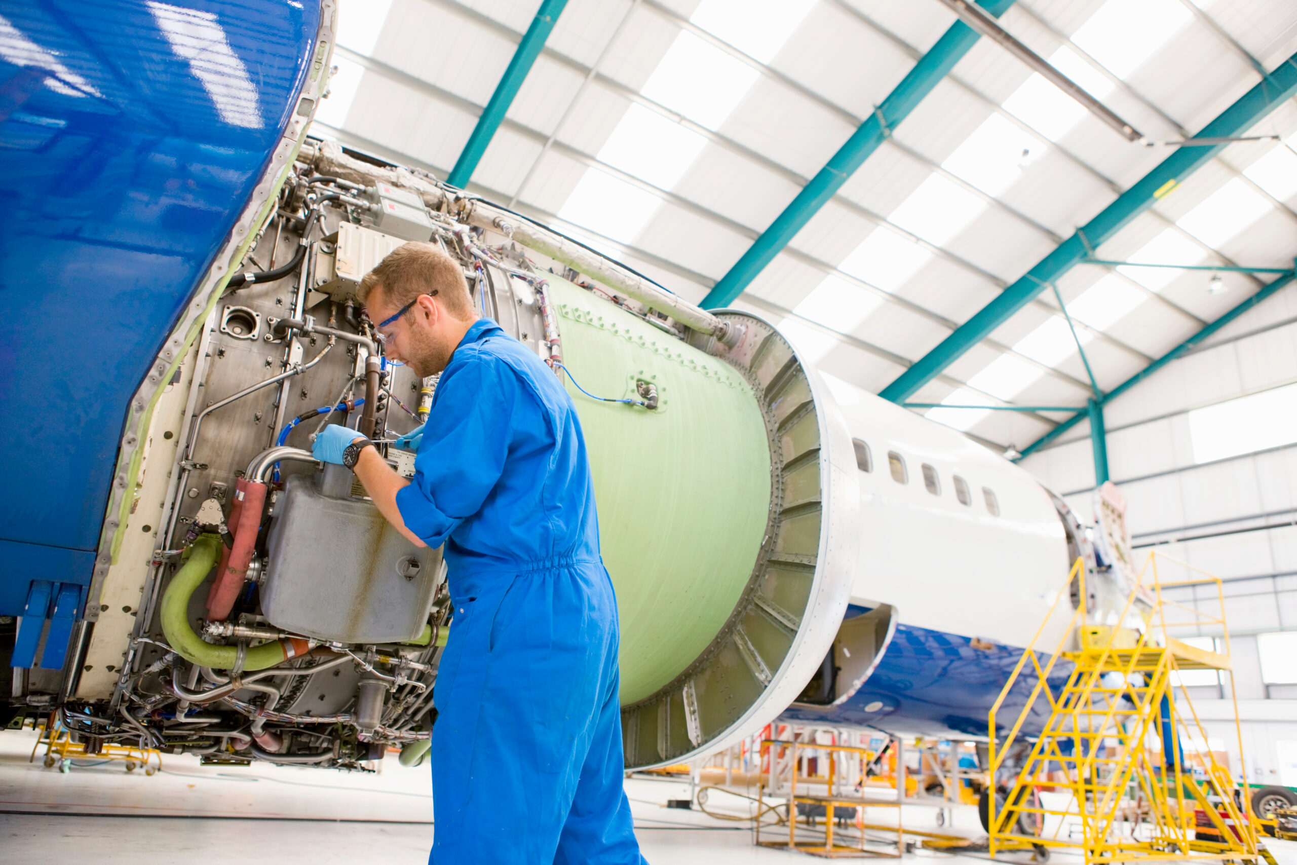 A skilled technician in blue overalls and safety glasses performing intricate maintenance and inspection work on the exposed engine section of a large aircraft in a hangar. This highlights the role of AI vision in the aerospace and defense industry for assembly verification, defect detection, and maintenance SOP compliance.