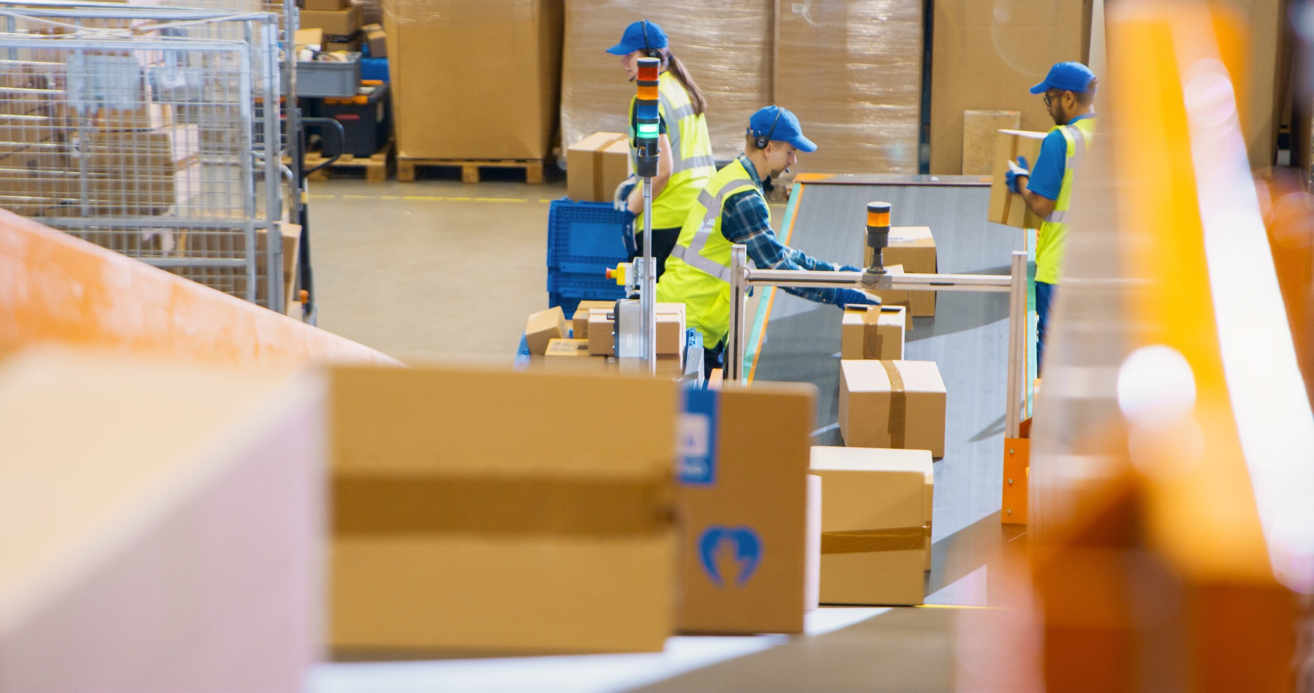 An active parcel sorting facility with warehouse operatives in high-visibility vests and blue caps manually handling packages on a network of conveyor belts under bright lighting. This illustrates how AI vision can enhance logistics operations through automated parcel scanning, handling, dimensioning, and quality control on busy sorting lines.