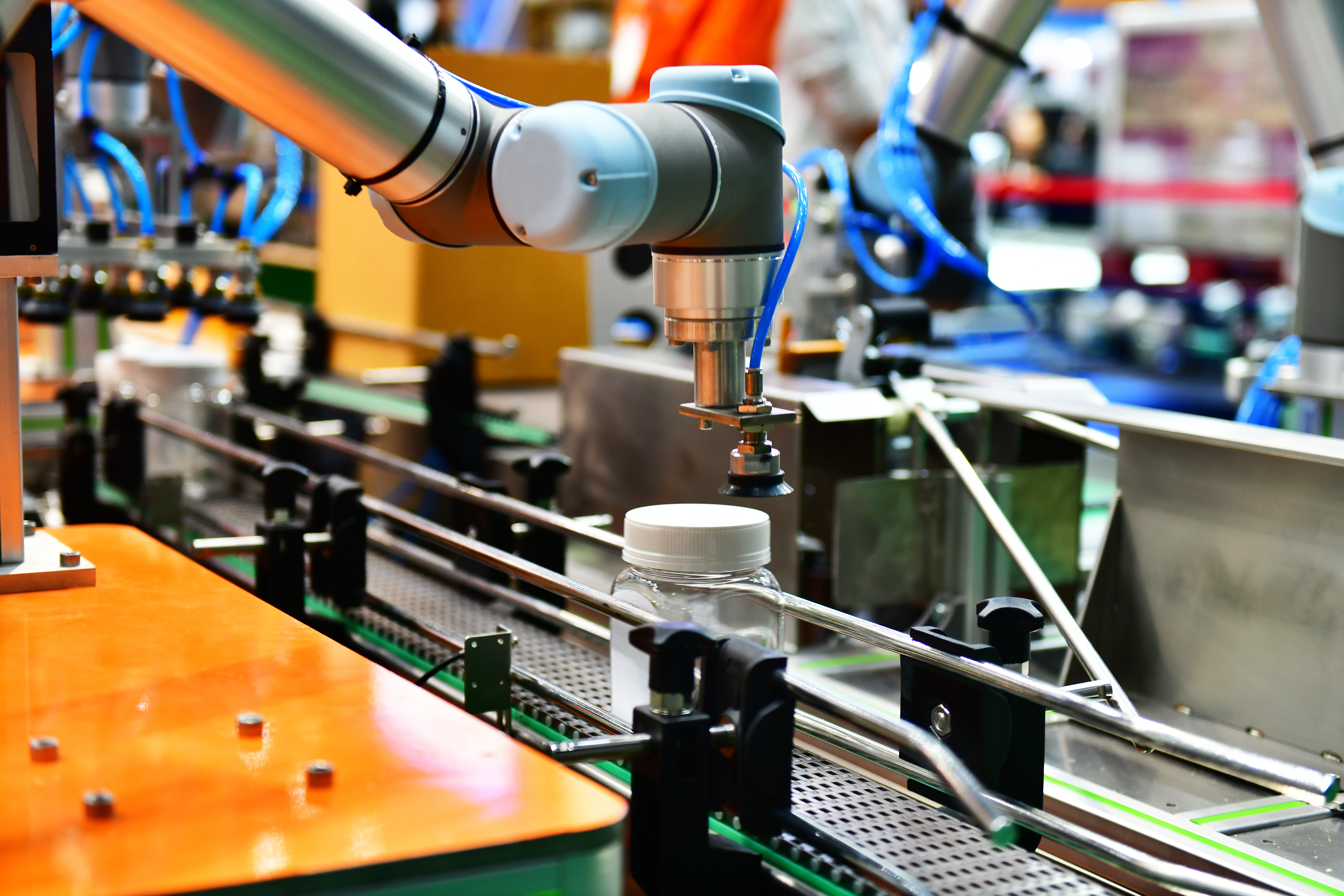 A close-up of a collaborative robotic arm accurately placing a white cap onto a clear jar on a conveyor belt in a brightly lit factory. This represents the application of AI vision in the manufacturing industry for robotic guidance, precise assembly, and quality inspection on the production line.