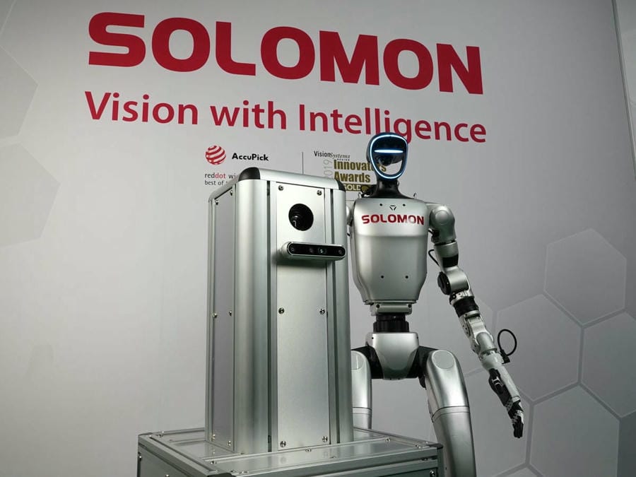Solomon humanoid robot with AI vision and an AMR in front of the Solomon logo and slogan in a warehouse.